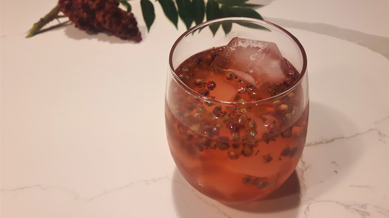 Glass of sumac lemonade with sumac berries and ice floating in the drink, with a cluster of sumac flowers in the background