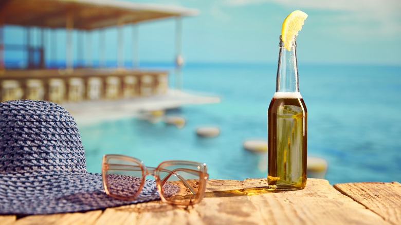 A bottle of beer on a pier with a lemon slice next to sunglasses and a hat
