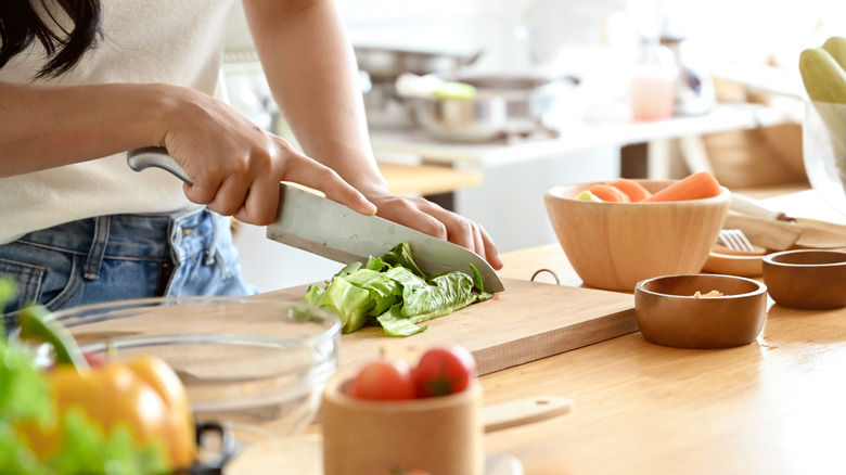 A person preparing vegetables on a chopping board, with assorted produce in a bowl on the table