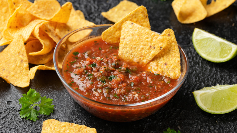 A bowl of tomato salsa surrounded by tortilla chips and limes
