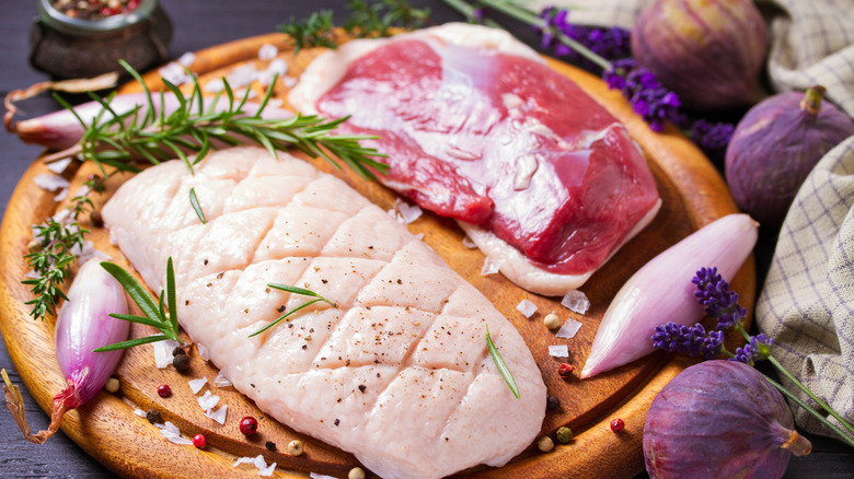 Two fatty pieces of raw duck on a cutting board surrounded by figs, shallots, herbs, and spices