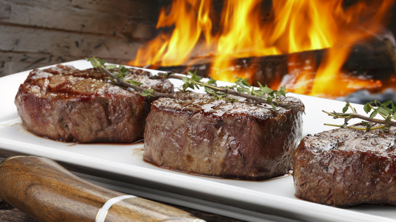 Three juicy steaks served on a white plate, with an open flame behind them