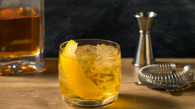 A rusty nail in a rocks glass is photographed with a bottle of liquor and bartending tools in the frame.