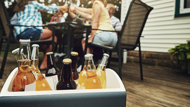 A cooler of beers on a patio with people toasting at a table in the background.