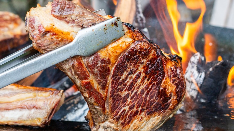 Steak being cooked to order on a grill held up by metal tongs.