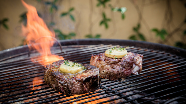 Steaks topped with compound butter cooking on a charcoal grill