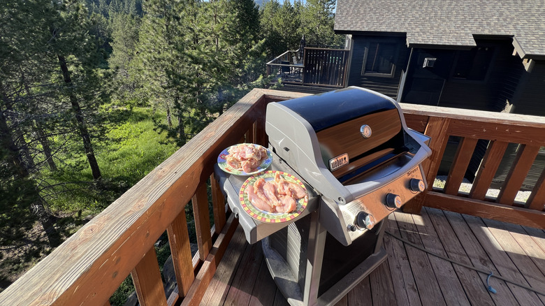 Gas grill with two plates of raw meat on the side tray