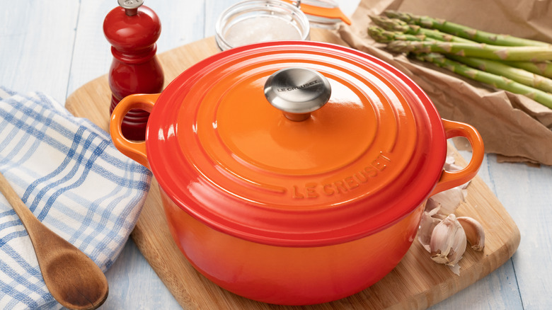 Orange Le Creuset Dutch oven on a wooden cutting board with garlic, a pepper grinder, and asparagus