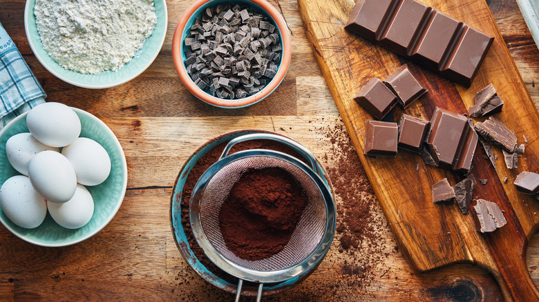 Wooden table with a bowl of eggs, a bowl of flour, cocoa powder in a seive, and a cutting board with pieces of chocolate