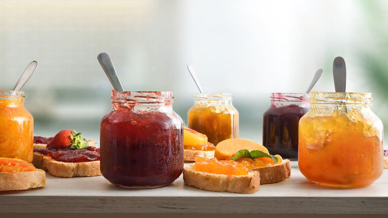 Open jars of preserves on a countertop alongside bread topped with preserves and fruit