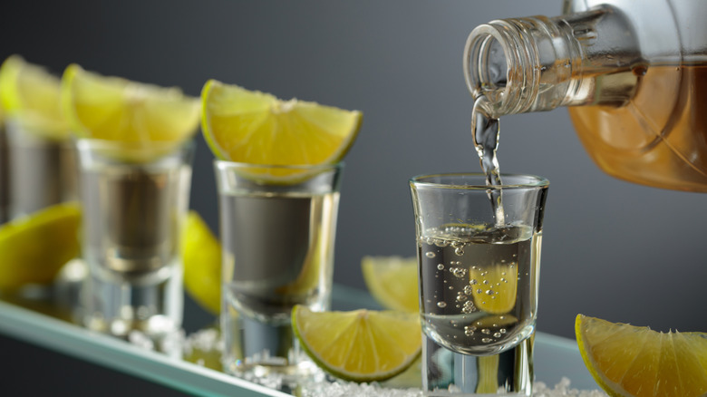 Tequila being poured from a bottle into a row of shot glasses with limes.
