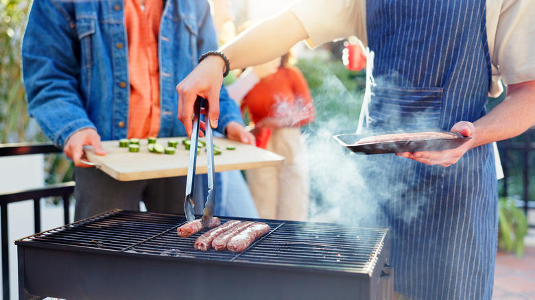 The person on the right wears an apron and is picking up sausages with tongs, while the other holds a wooden board with sliced cucumbers.