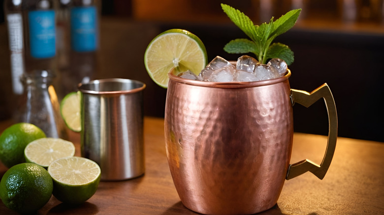 A mule in a copper cup sits on a bar with limes and barware nearbuy.
