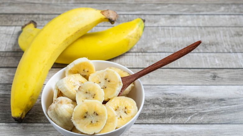 Two whole yellow bananas sitting next to a bowl of sliced bananas