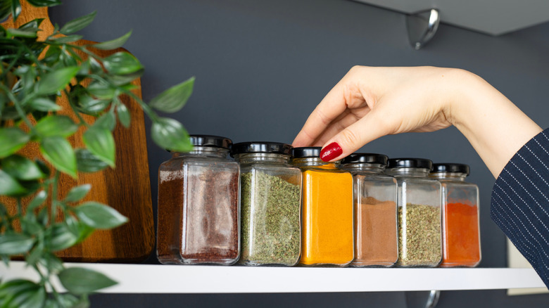 Hand reaching out to grab a jar of spice on shelf next to cutting board and plant.