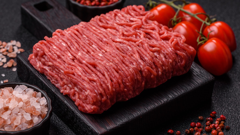 Raw ground beef on wooden cutting board with rock salt and vine tomatoes beside it.
