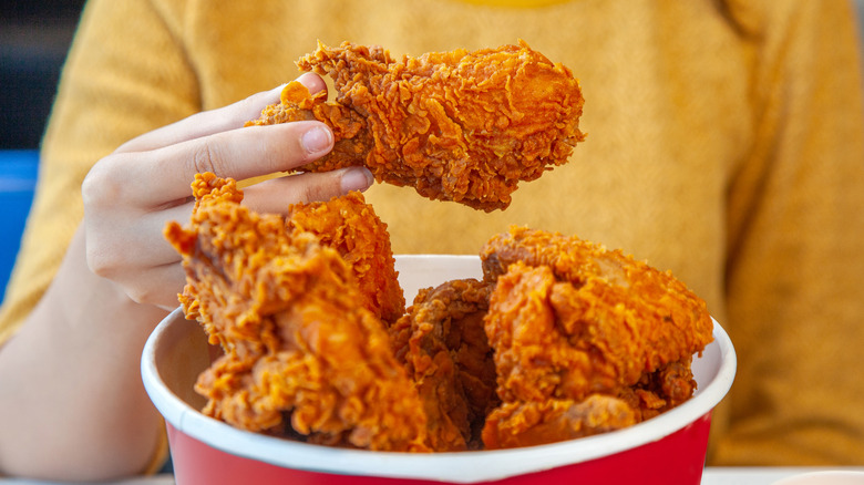 A hand holding a piece of fried chicken over a bucket of fried chicken.