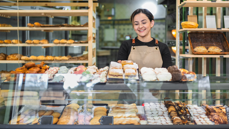A baker in a black shirt and tan apron standing behind a glass pastry case with assorted pastries and baked goods.