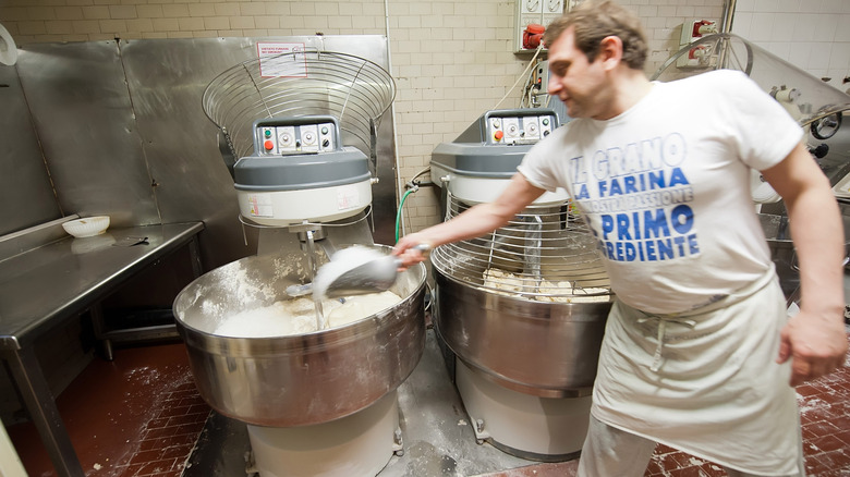 A commercial baker in a white and blue T-shirt adds ingredients to a large stand mixer.