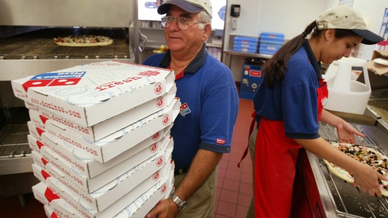 Two Domino's employees in the kitchen of a Domino's store with one carrying pizza boxes and the other making a pizza ready for the oven.