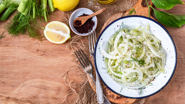 A raw, shaved fennel salad, topped with fennel fronds, surrounded by fennel stalks, sliced lemon, and spices