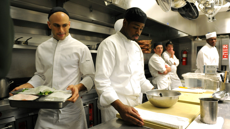 Assistant White House chef Sam Kass, left, and other members of the kitchen staff prepare food for the first formal dinner of Barack Obama's presidency, the National Governors Association dinner, in Washington, D.C. on February 22, 2009. Kass carries a tray of food to the counter, where another cook stands at a cutting board with a large mixing bowl; three other cooks look on in the background.