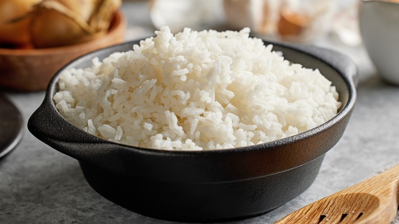 Plain white rice piled in a cast iron bowl next to a wooden spoon on a gray countertop.