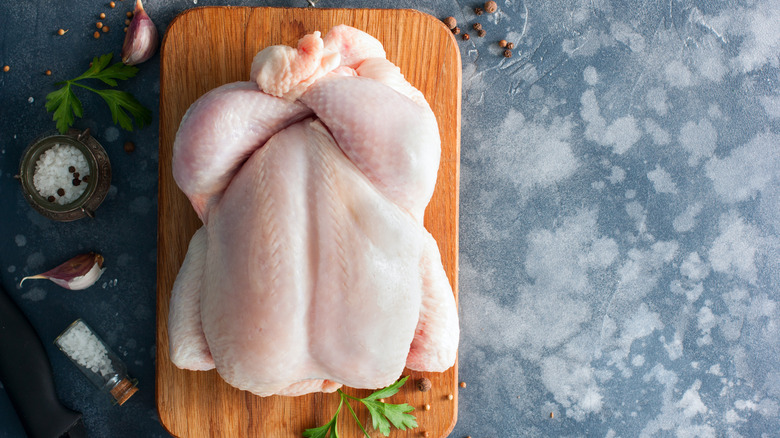 A whole raw chicken on a wooden cutting board next to spices and herbs