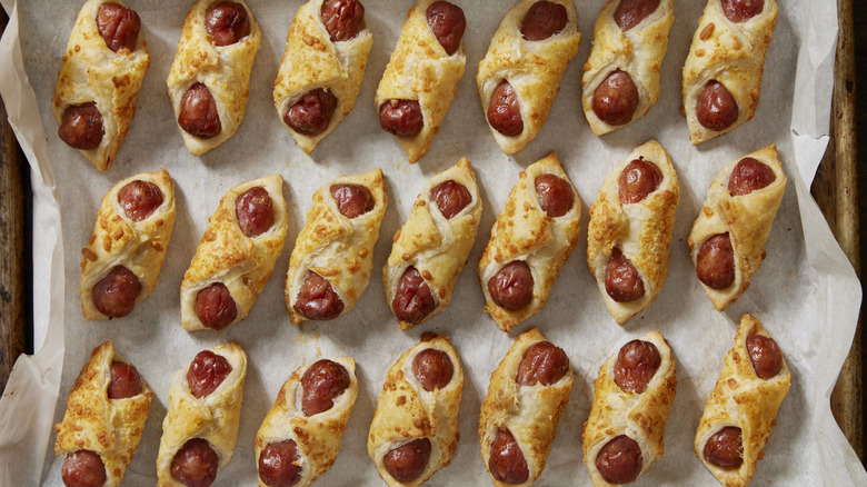 Rows of pigs in a blanket top a parchment-lined baking sheet.