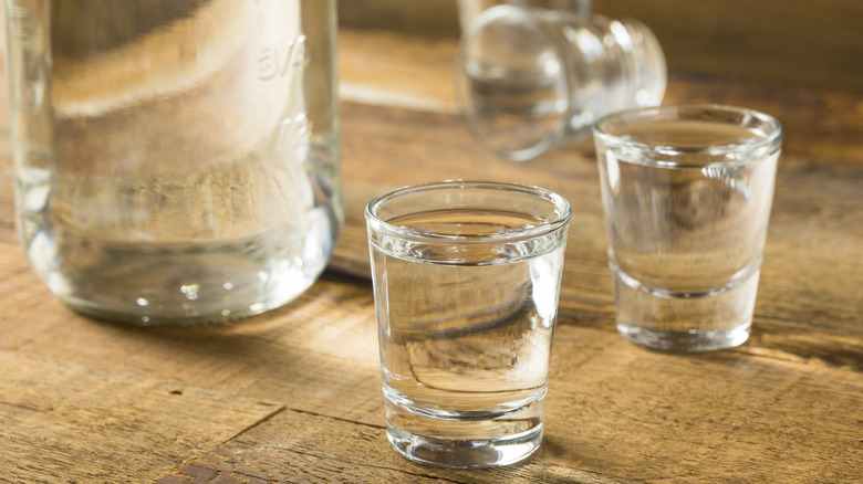 Shot glasses with a clear spirit in them and the bottle nearby, all on a wooden table.