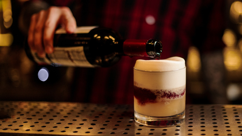 A bartender pours red wine into a double old fashioned glass to top off a foamy New York Sour cocktail