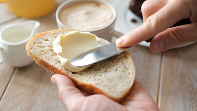 Hand using butter knife to spread butter on slice of white Italian bread with coffee and cream in background on wood surface.