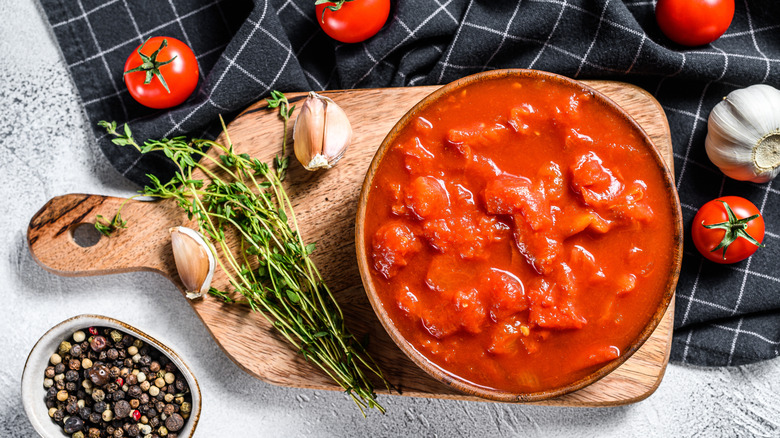 Canned, chopped Roma tomatoes in a bowl on a cutting board surrounded by fresh thyme, garlic cloves, fresh cherry tomatoes, and a bowl of black peppercorns.