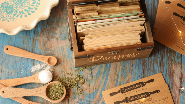 A box of recipes next to spoons filled with ingredients on a blue wooden table