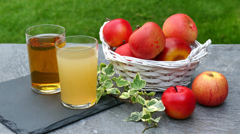 Glasses of clear and cloudy apple juice next to a basket of whole red apples on a gray surface