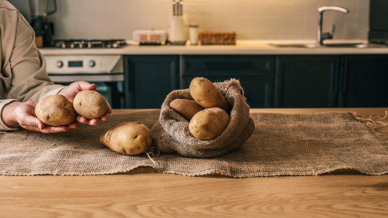 Hands holding and arranging fresh potatoes in a burlap sack on a wooden table in a cozy kitchen. Warm lighting and rustic atmosphere.