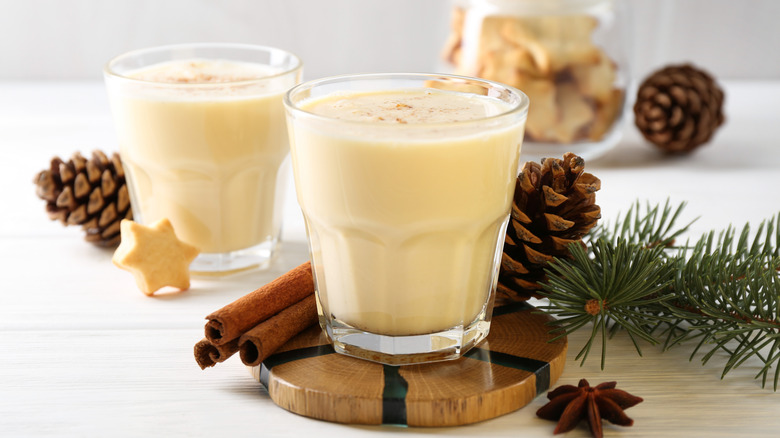 Two glasses of eggnog near pinecones and cinnamon sticks on a white background