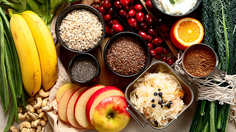 Overhead view of various foods on a table, including bananas, cashews, a sliced apple, cranberries, an orange, bowls of grains, and more