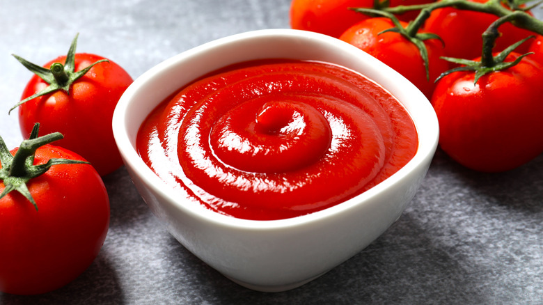 White ramekin of ketchup on gray slate table surrounded by tomatoes.