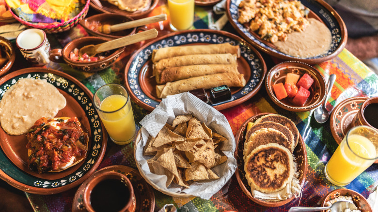 Table of Mexican food with tortilla chips, taquitos, tacos, fruit, juice, and more