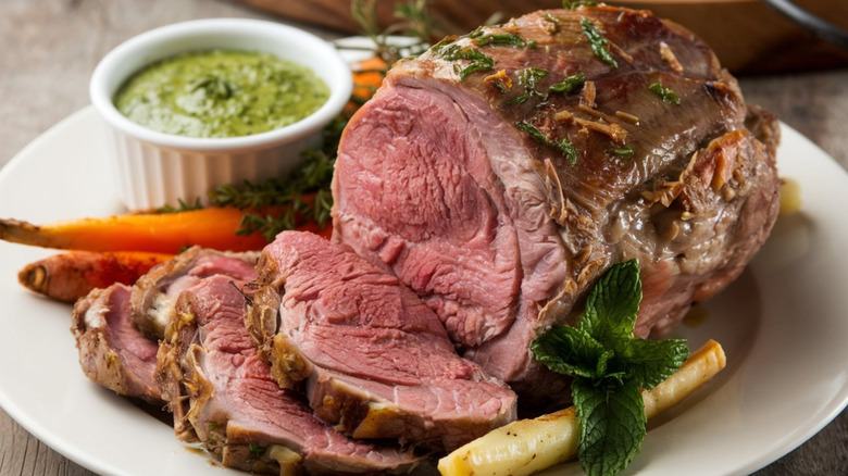 A lamb roast on a platter alongside vegetables and sauce