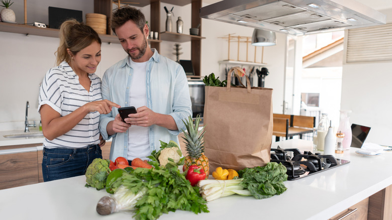 Couple using a phone in a kitchen with a partly unloaded bag of groceries on the island