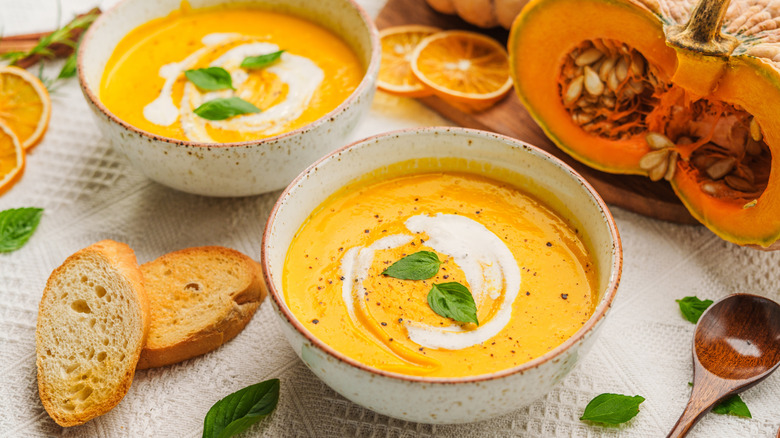 Two bowls of hot cream of pumpkin soup with a milky swirl in the middle, topped with basil leaves, the bowls surrounded by crunchy slices of French bread, lemon wheels, and a fresh pumpkin cut in half