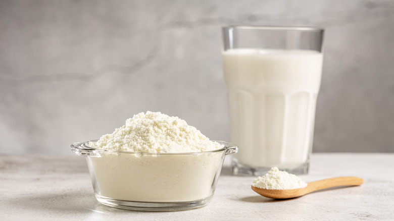 A small bowl of heaped powdered milk with a wooden spoonful of powdered milk next to it, a glass of mixed powdered milk behind them on a grey countertop