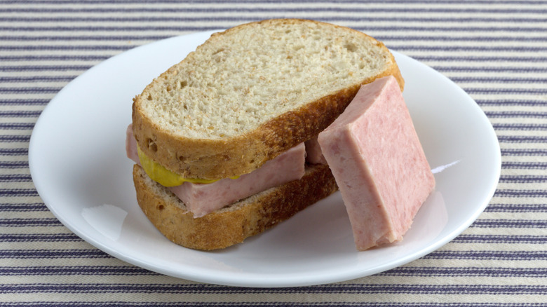 A canned pork sandwich with mustard, along with an extra chunk of meat on the side, sits atop a plate on a striped tablecloth.