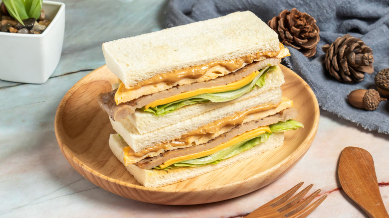 A sandwich consisting of peanut butter, egg, pork, cheese, and lettuce on white bread sits atop a wooden plate on a table.
