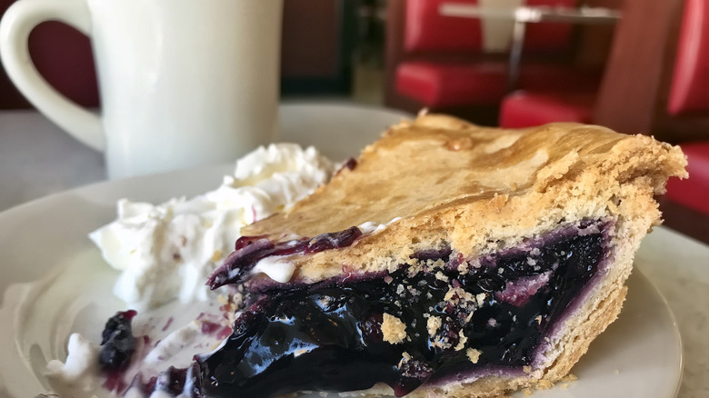 A slice of blueberry pie with cream sits on a plate next to a cup of coffee at a diner table
