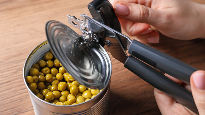 Somebody opens a tin of green peas with a can opener at wooden table
