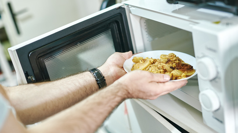 A person placing a plate of casserole leftovers in a microwave.