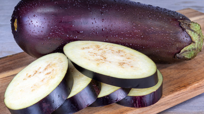 A whole eggplant with sliced pieces of eggplant sitting in front of it on a wooden cutting board.